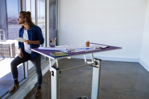 Mężczyzna patrzy przez okno obok biurka z planami, które ma stelaze regulowane.