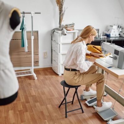 Kobieta szyje przy maszynie w pracowni, gdzie w tle widoczne są stelaze regulowane recznie.