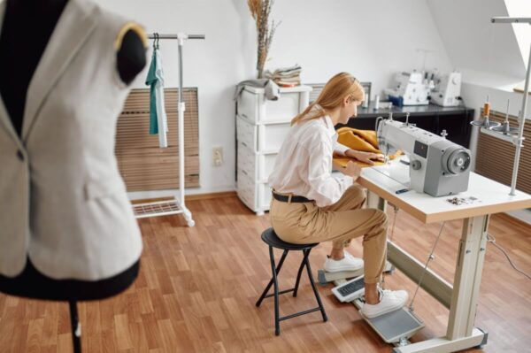 Kobieta szyje przy maszynie w pracowni, gdzie w tle widoczne są stelaze regulowane recznie.