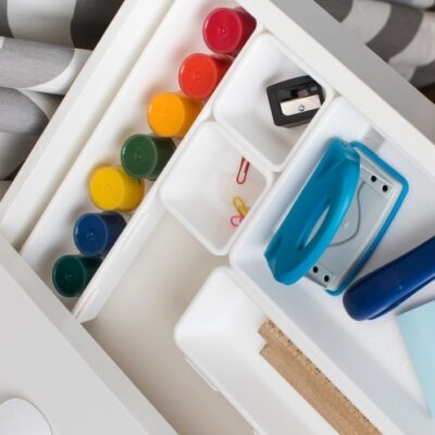 W szufladzie metalowej poukładane są kolorowe farby i przybory biurowe.